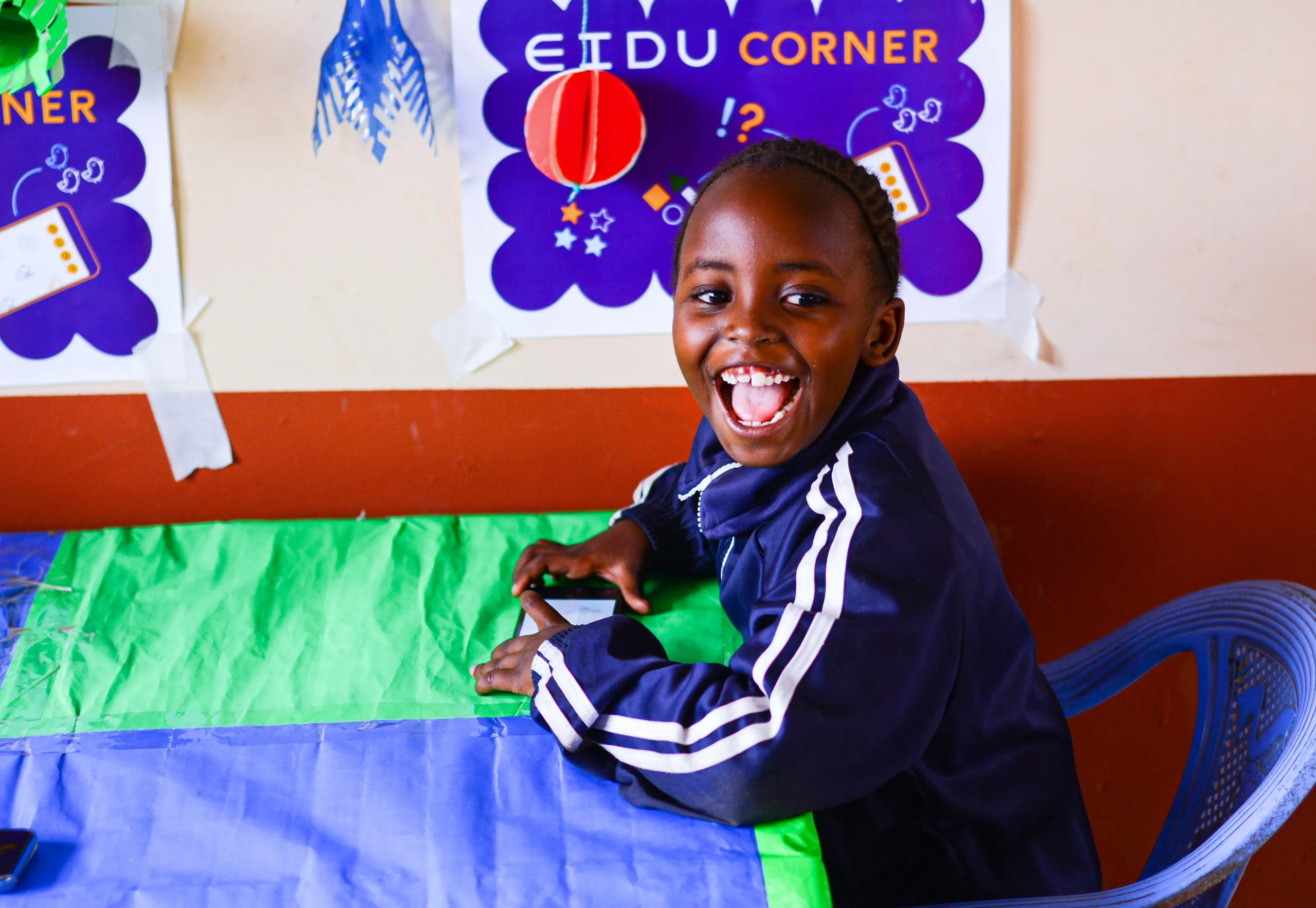 Smiling girl at a desk with an EIDU phone, in front of an EIDU Corner poster in her classroom