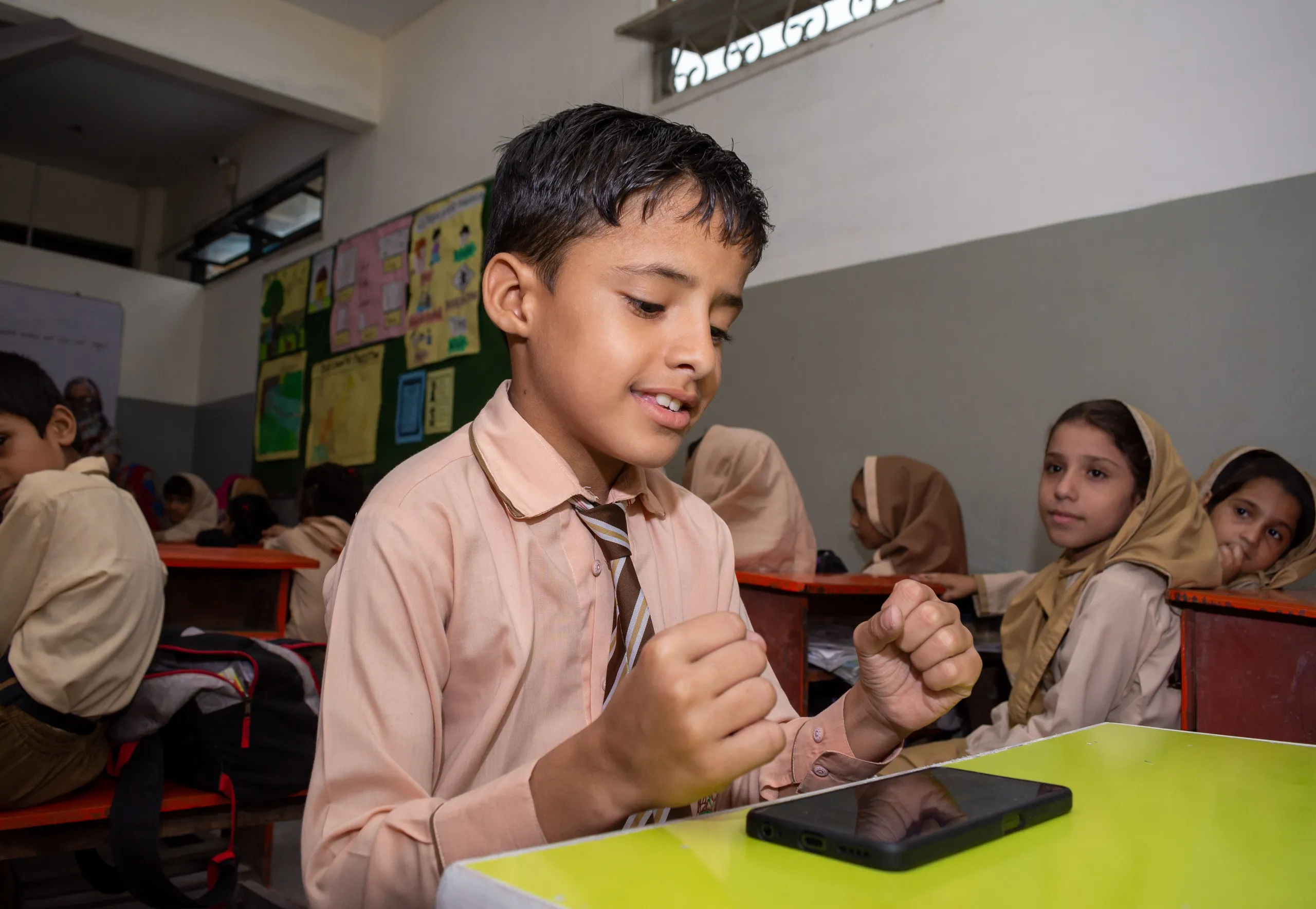 Smiling boy in a school uniform with an EIDU phone on his desk as classmates look on