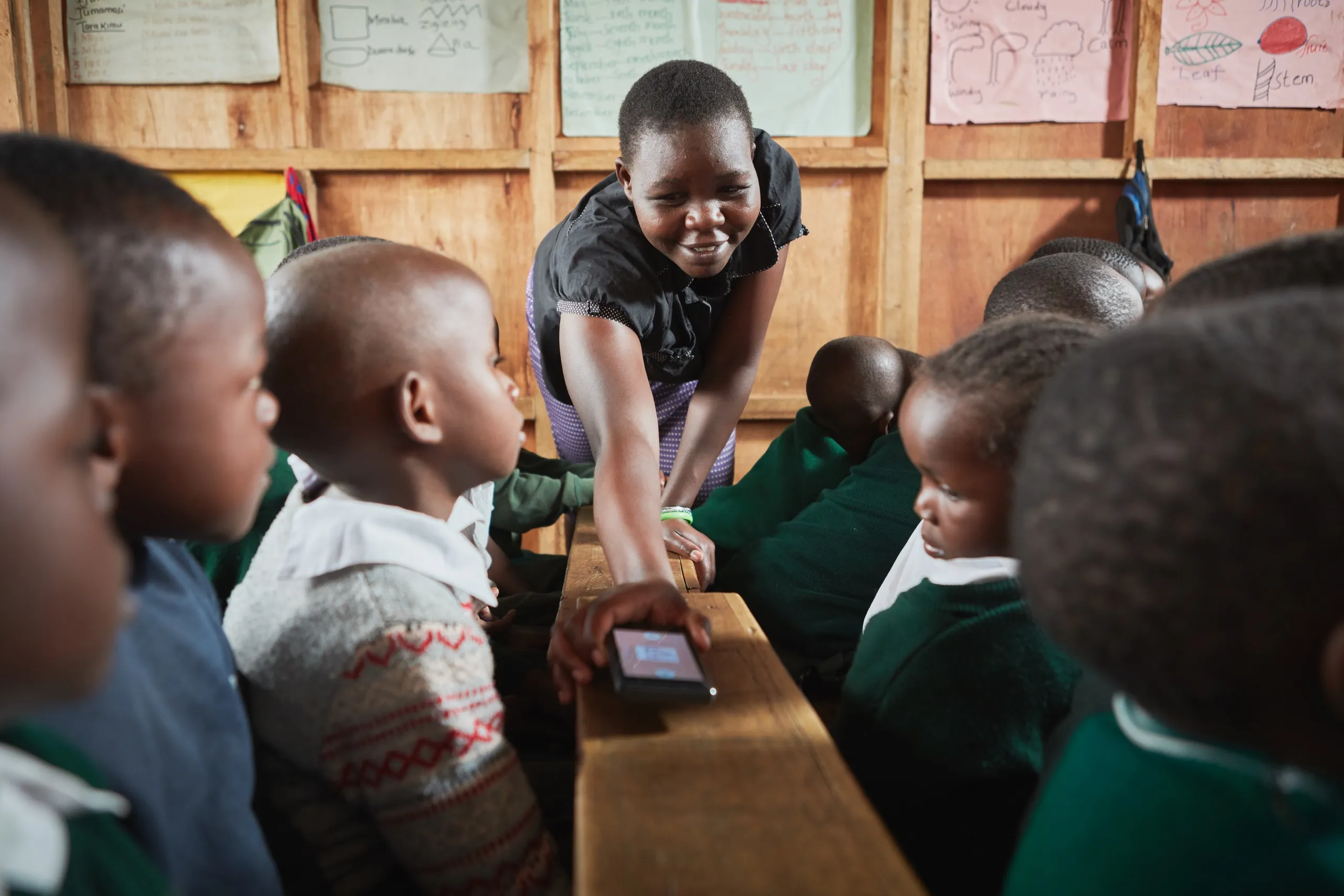 Teacher handing a phone to pre-primary pupils gathered around a classroom bench