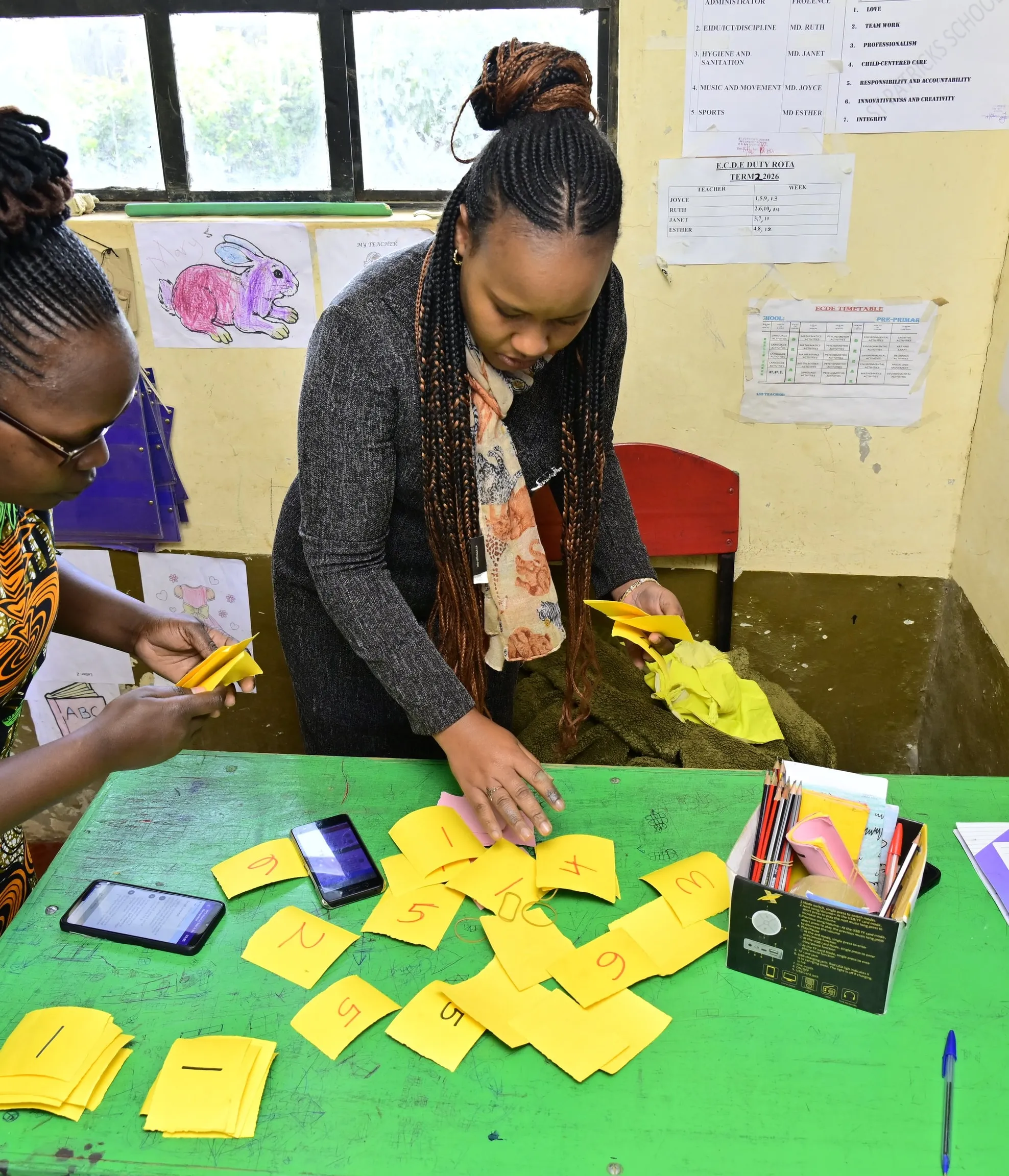 Two teachers in a Kenyan pre-primary classroom sorting handmade number cards next to EIDU phones