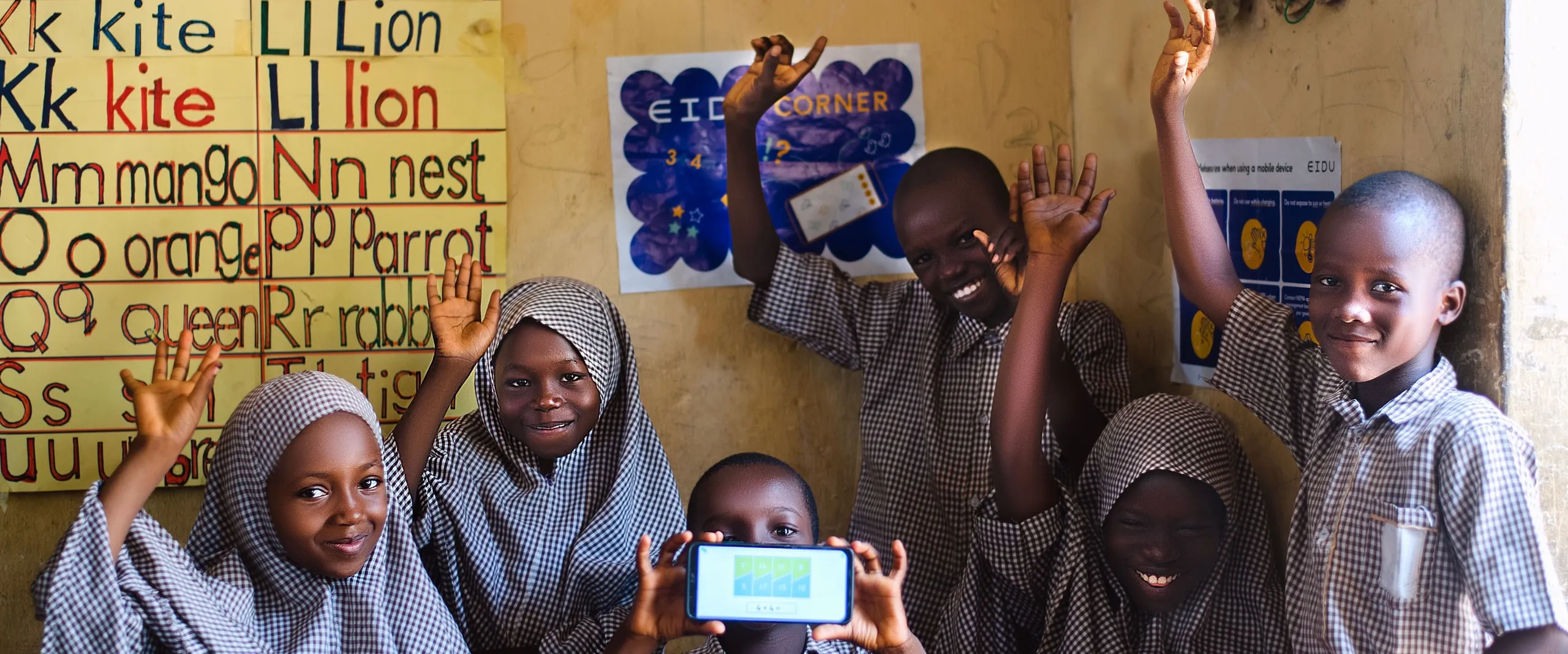 Six learners cheering with their hands up in front of an alphabet chart, one holding up a phone, in Adamawa State, Nigeria