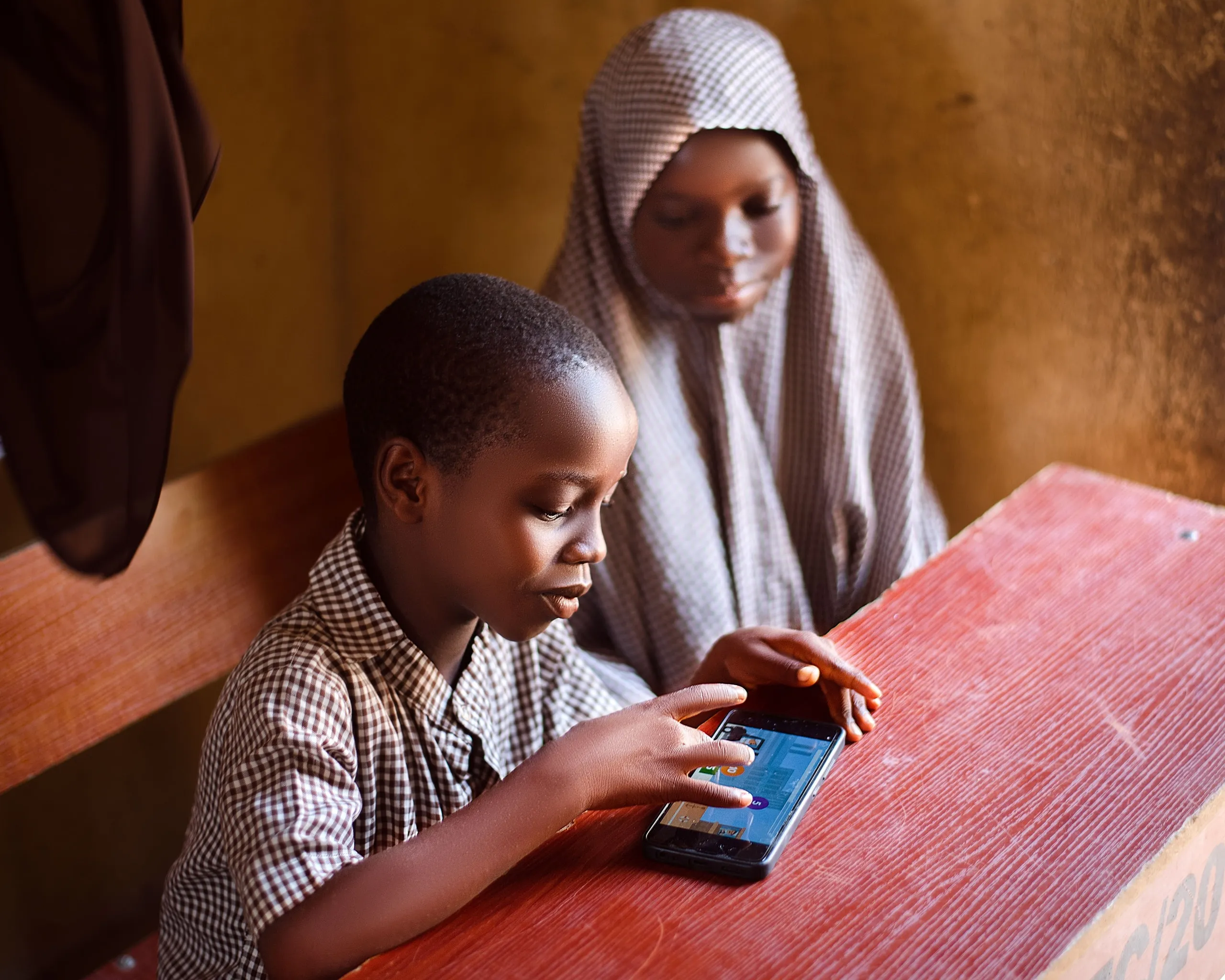 Two learners doing a lesson on a phone at a wooden desk in Adamawa State, Nigeria