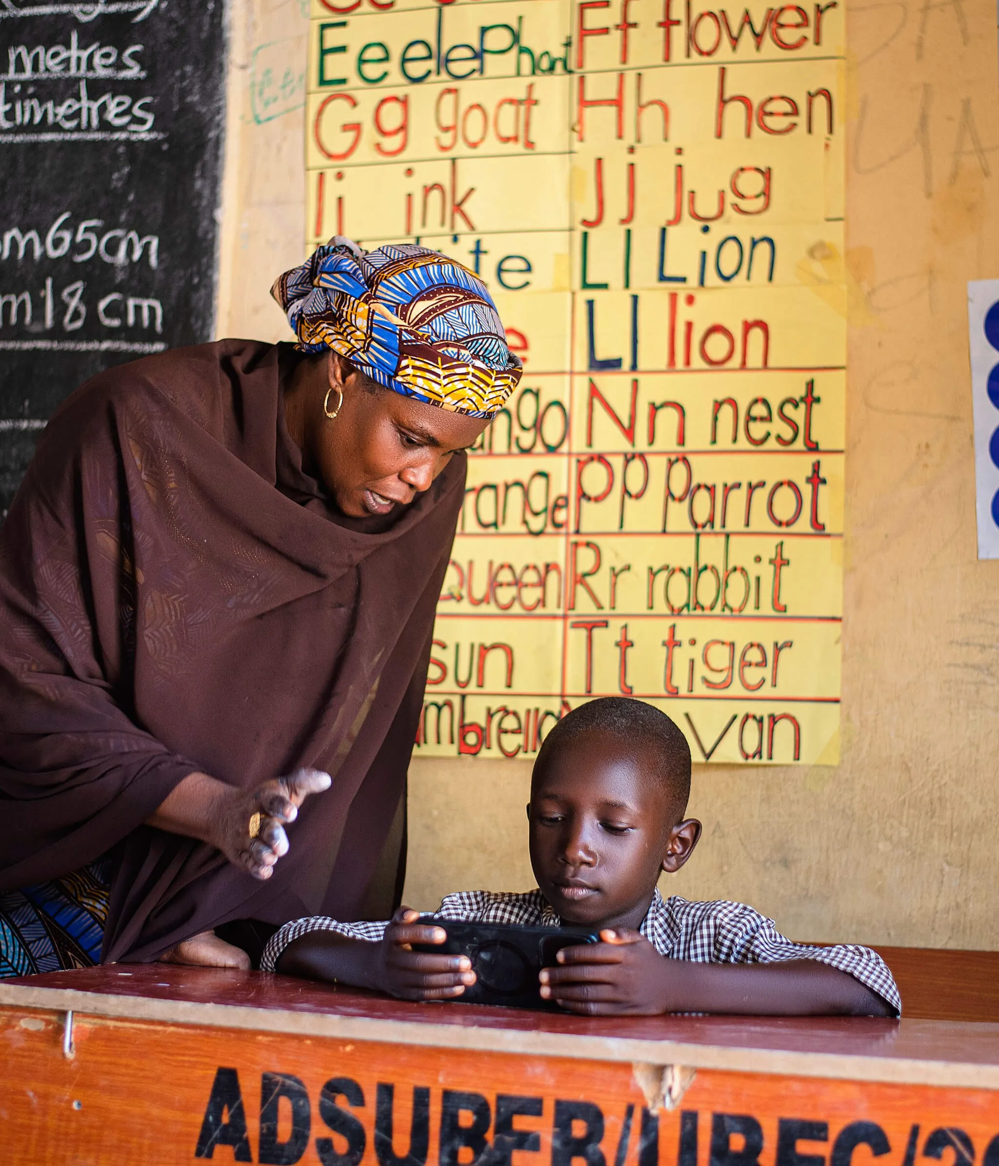 Teacher guiding a boy through a lesson on an EIDU phone in front of an alphabet chart in a Nigerian classroom