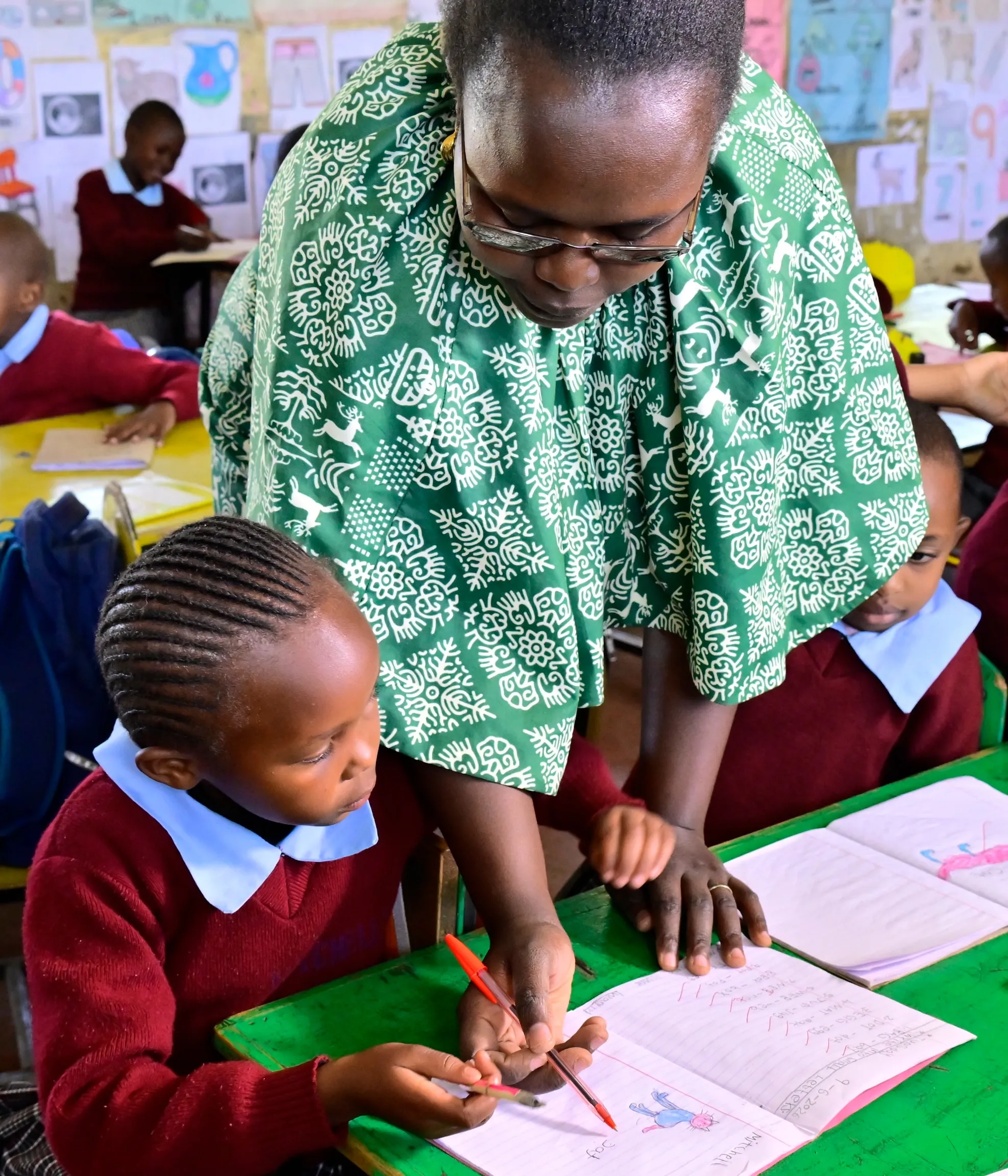 A teacher leaning over a girl to guide her writing in a colourful classroom in Nakuru County, Kenya