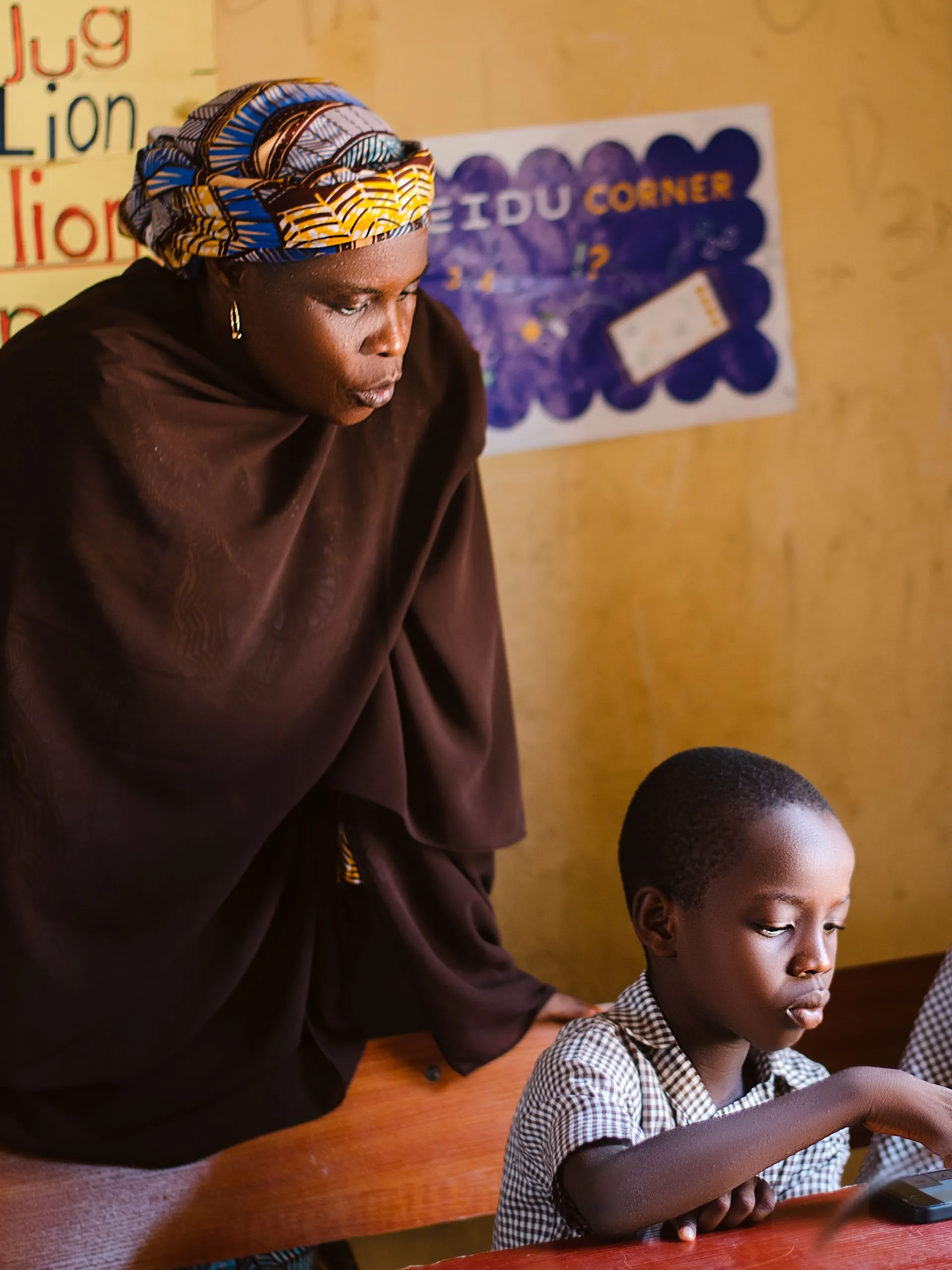 A teacher in a patterned headwrap watching a boy do a lesson on a phone in Adamawa State, Nigeria