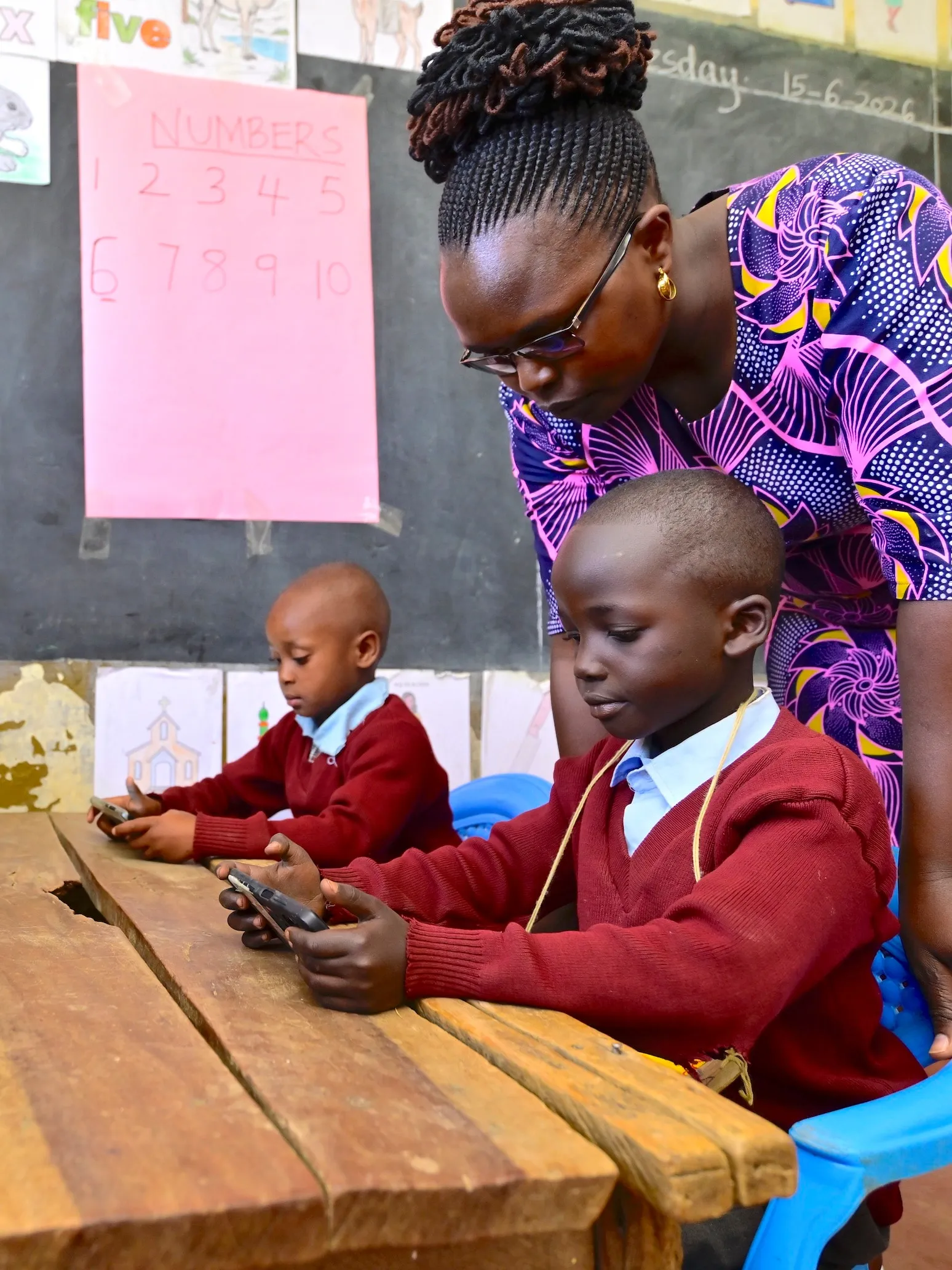 A teacher leaning over two boys using phones at the digital corner of their classroom in Nakuru County, Kenya