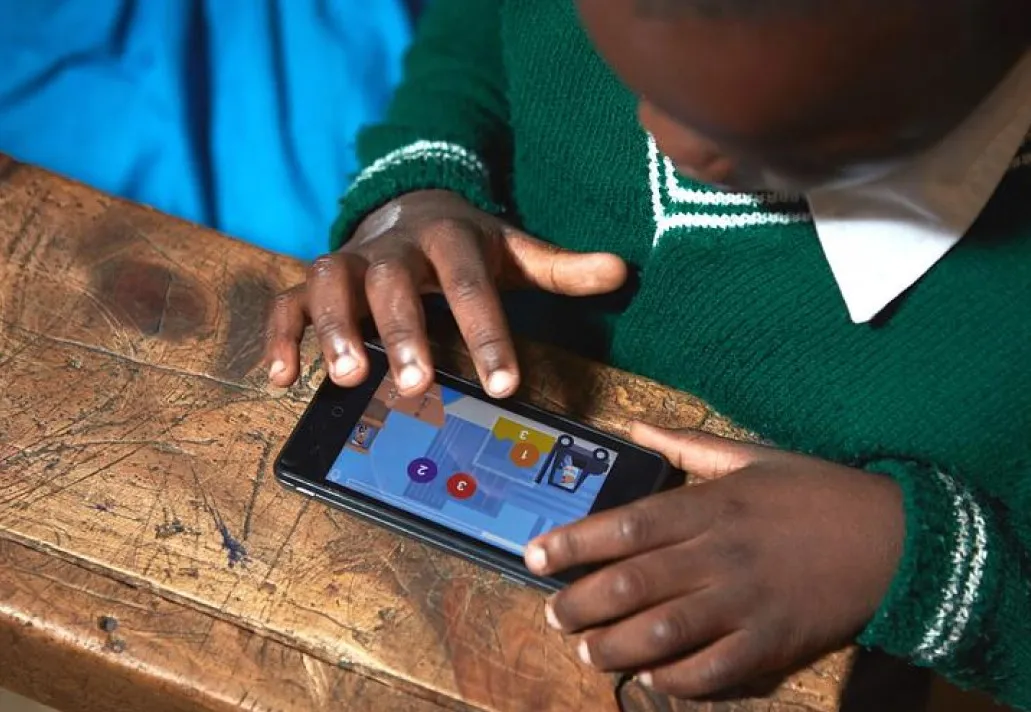 Child in a green school jumper playing a counting game on a phone at a wooden desk